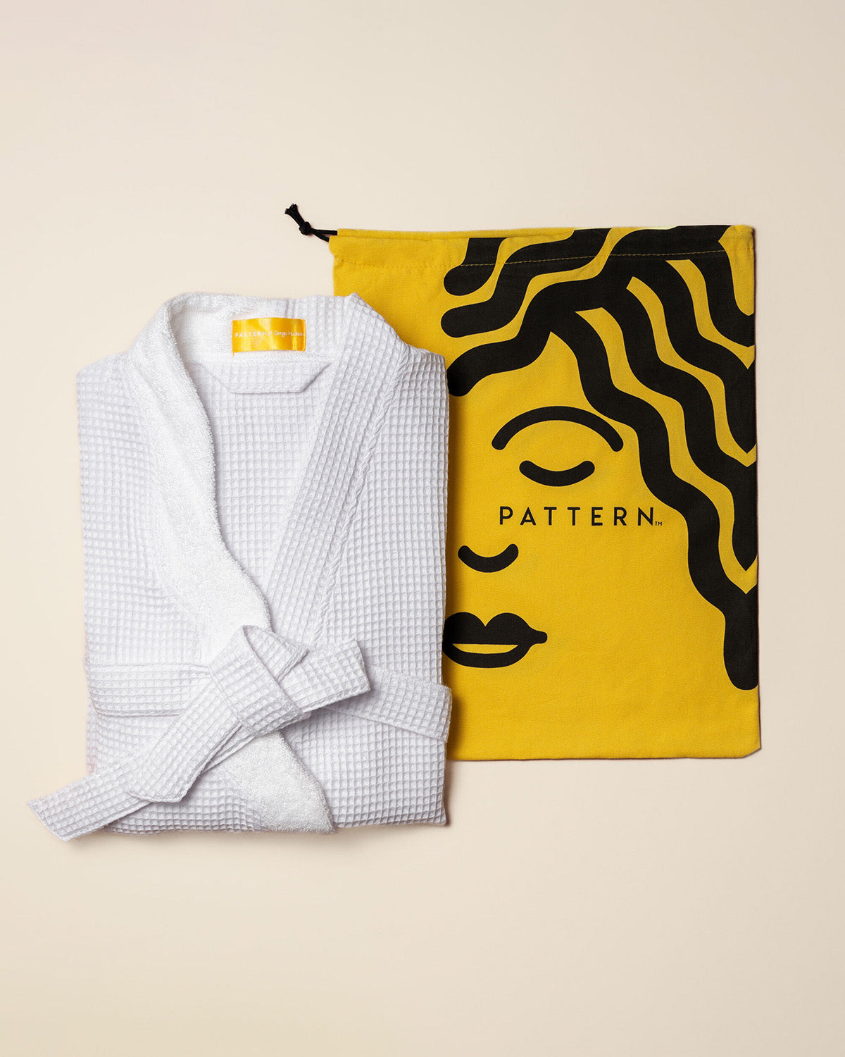 PATTERN x SERGIO HUDSON Curl-Conscious Robe - White waffle-textured cotton robe next to a mustard yellow pouch featuring black abstract face and hair design.
