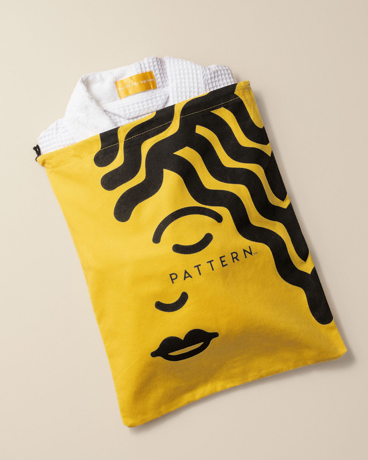 PATTERN x SERGIO HUDSON Curl-Conscious Robe - Yellow fabric pouch with black abstract face design and PATTERN logo, holding a white textured robe.
