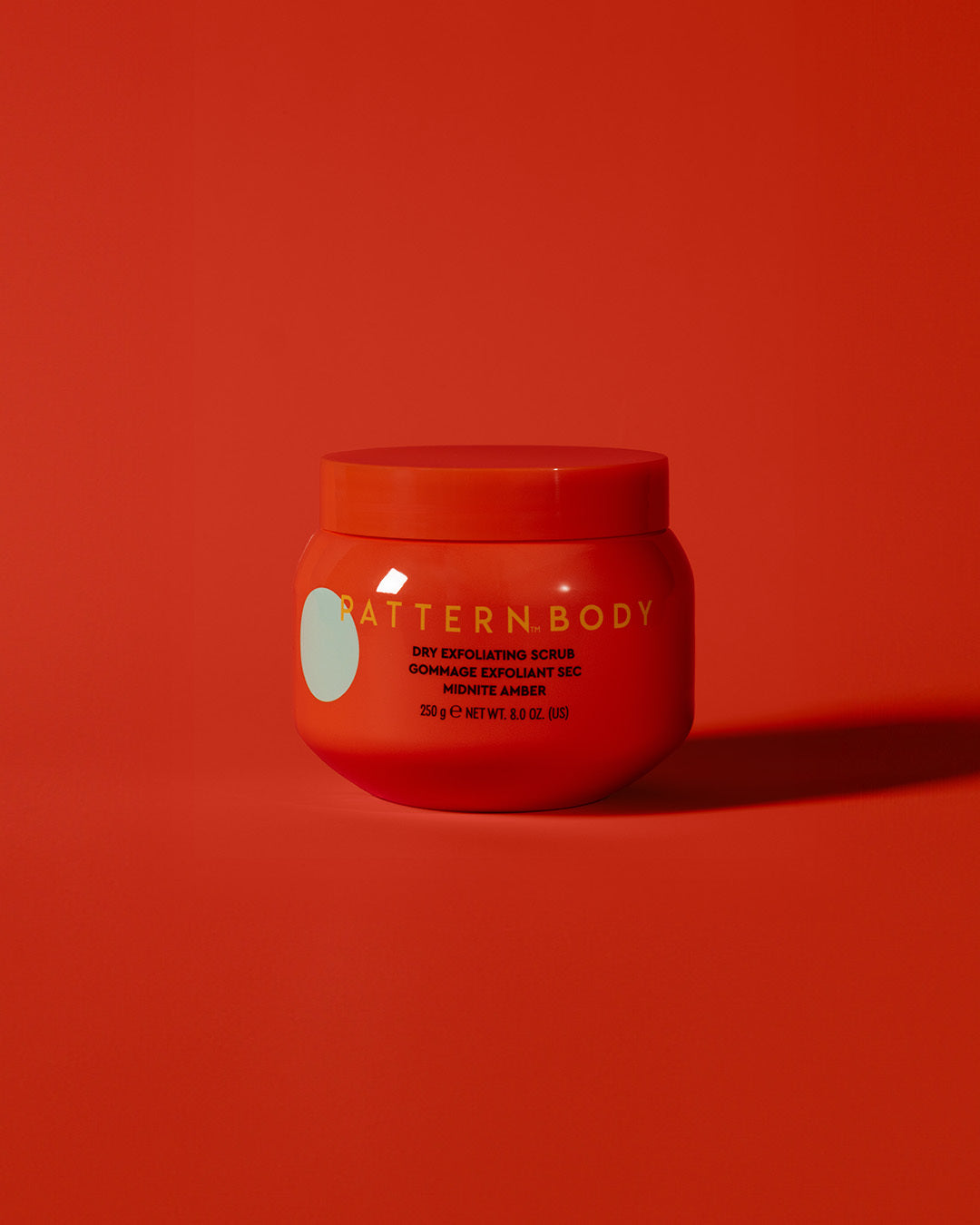 Dry Exfoliating Body Scrub - Red jar of PATTERN Beauty by Tracee Ellis Ross Dry Exfoliating Body Scrub with minimalist label on vibrant orange background.