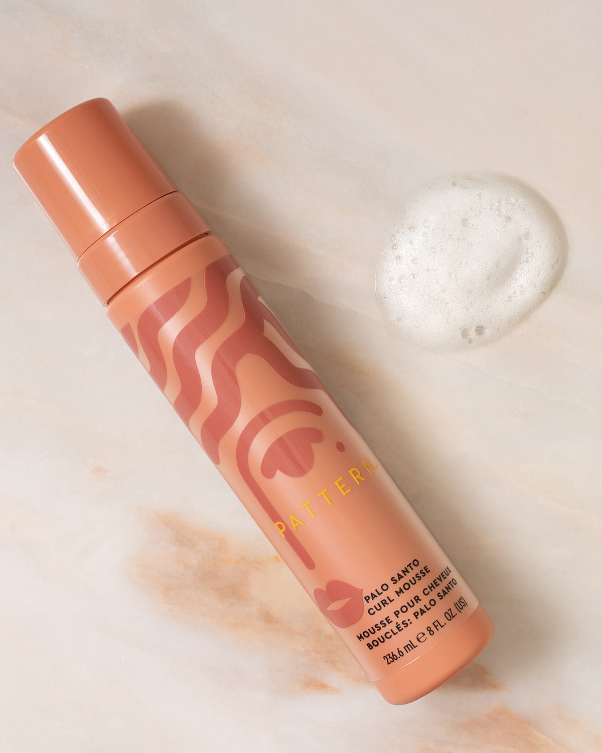 Palo Santo Curl Mousse - Matte peach cylindrical bottle with wavy pattern and foam next to it for PATTERN Beauty Palo Santo Curl Mousse.