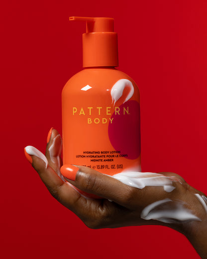 BODY-TEST GWP variant test - Orange pump bottle of PATTERN Beauty by Tracee Ellis Ross body lotion held with white cream on hand against red background.