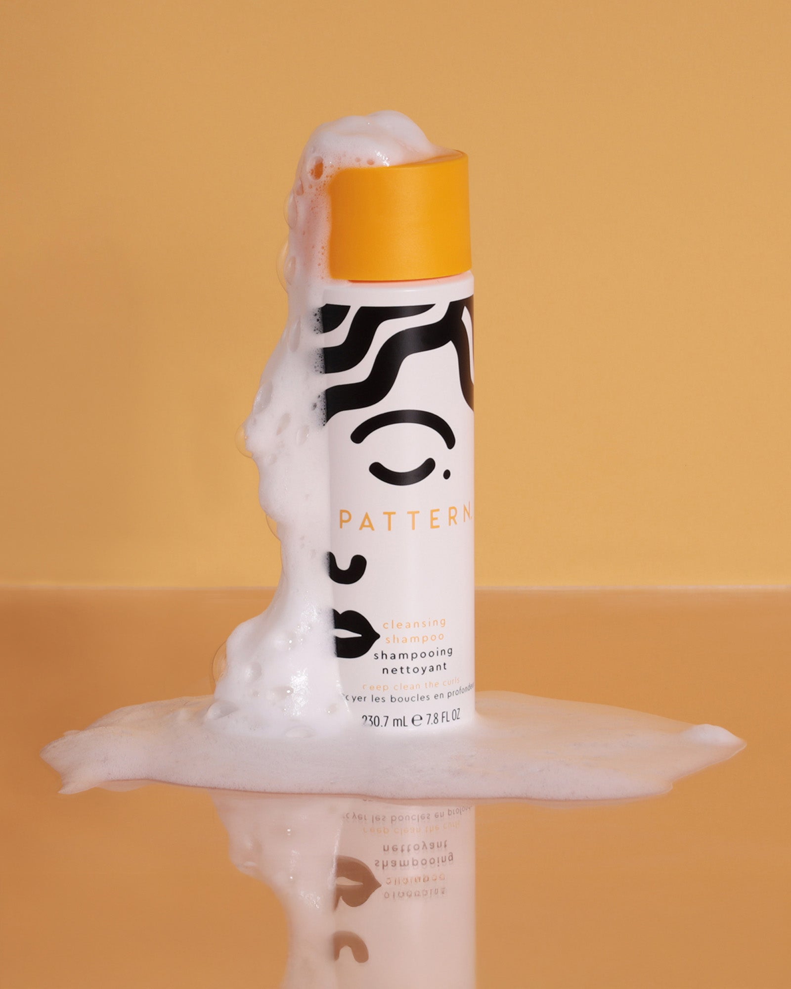 Cleansing Shampoo - White bottle with black abstract patterns and an orange cap, frothy shampoo overflowing from the top.