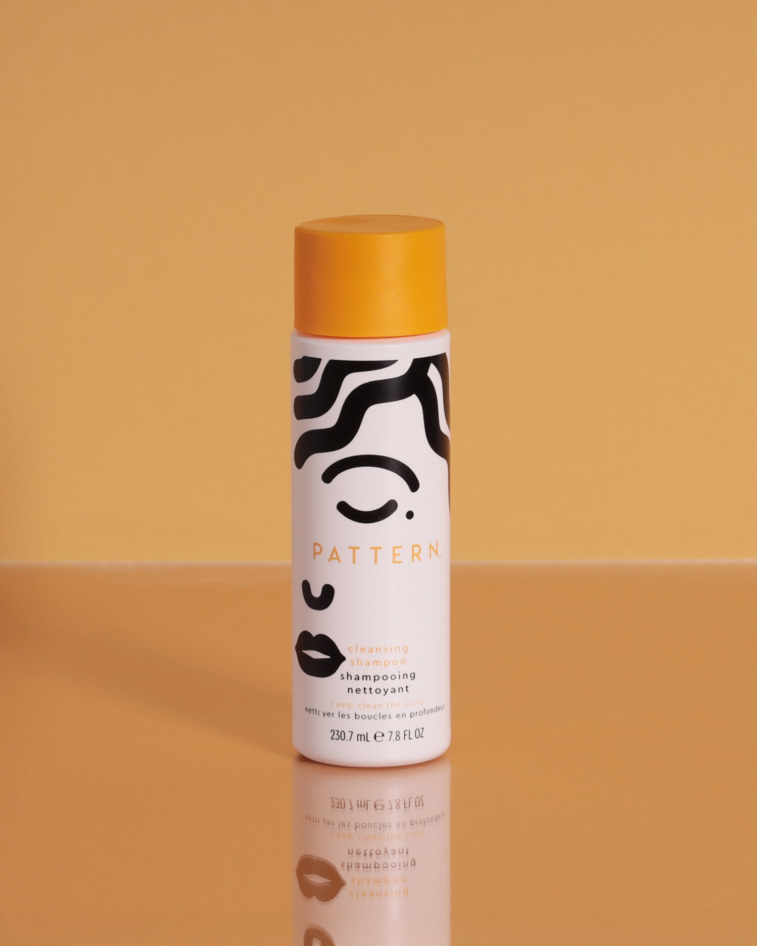 Cleansing Shampoo - White cylindrical bottle with black abstract face design and orange cap labeled PATTERN Beauty Cleansing Shampoo.
