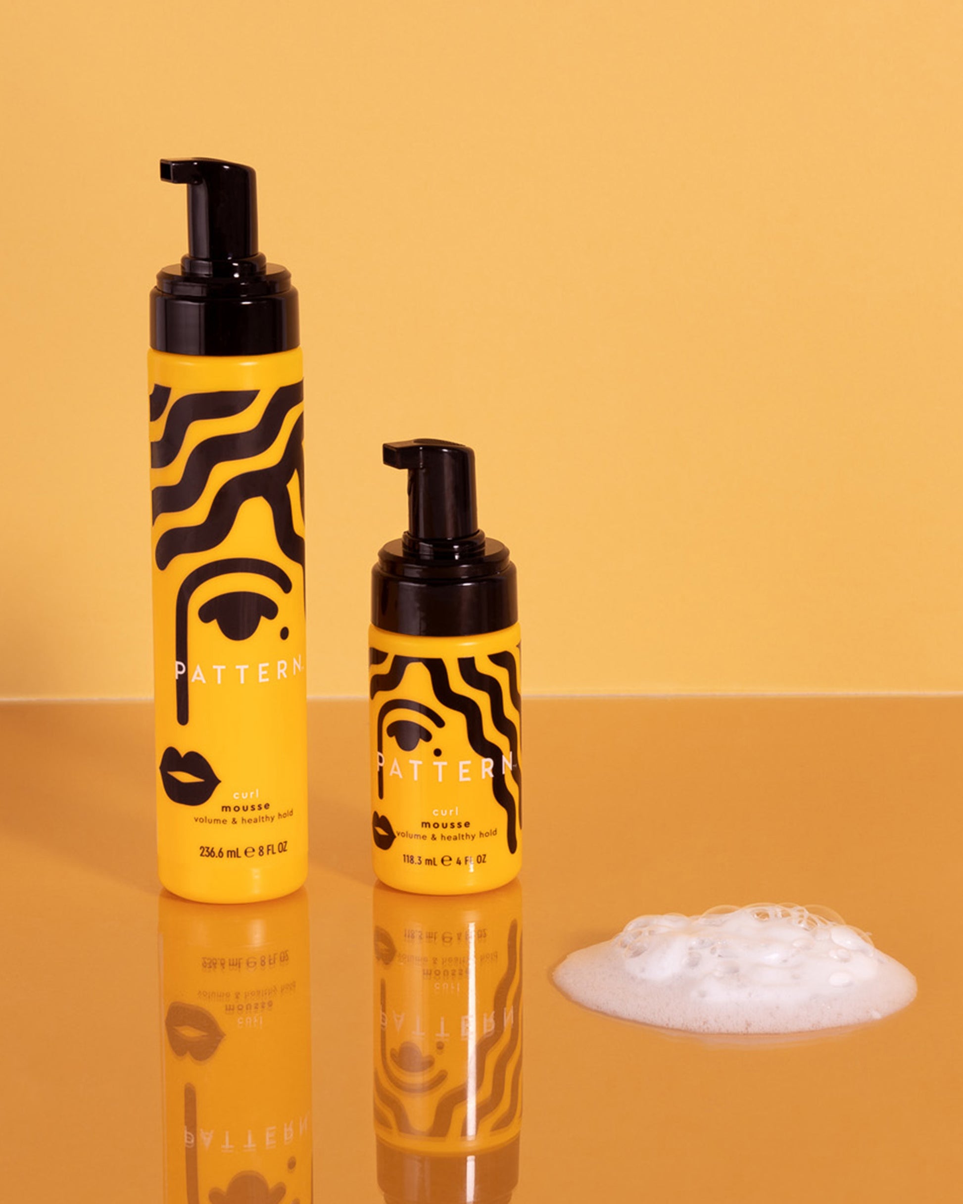 Curl Mousse - Two yellow bottles of PATTERN Beauty Curl Mousse by Tracee Ellis Ross with black pump tops and bold black wavy stripes, foam