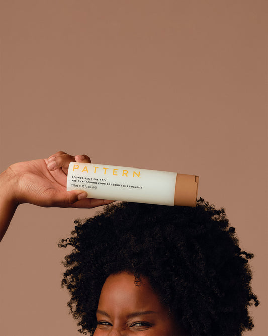 Bounce Back Pre-Poo - Hand holding PATTERN Beauty Bounce Back Pre-Poo tube above natural curly hair against beige background.
