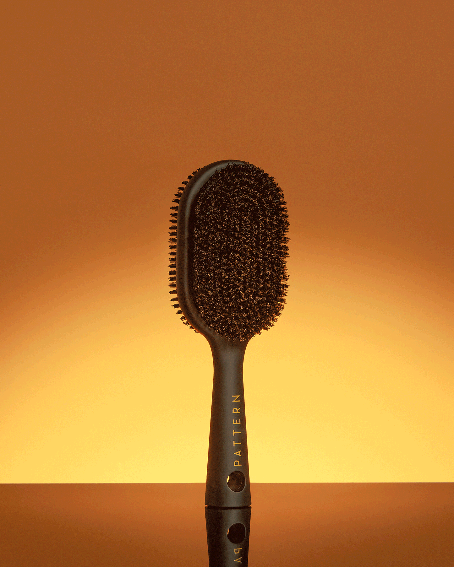 Double-Sided Bristle Brush - PATTERN Beauty by Tracee Ellis Ross double-sided black bristle brush with ergonomic handle against warm gradient background.