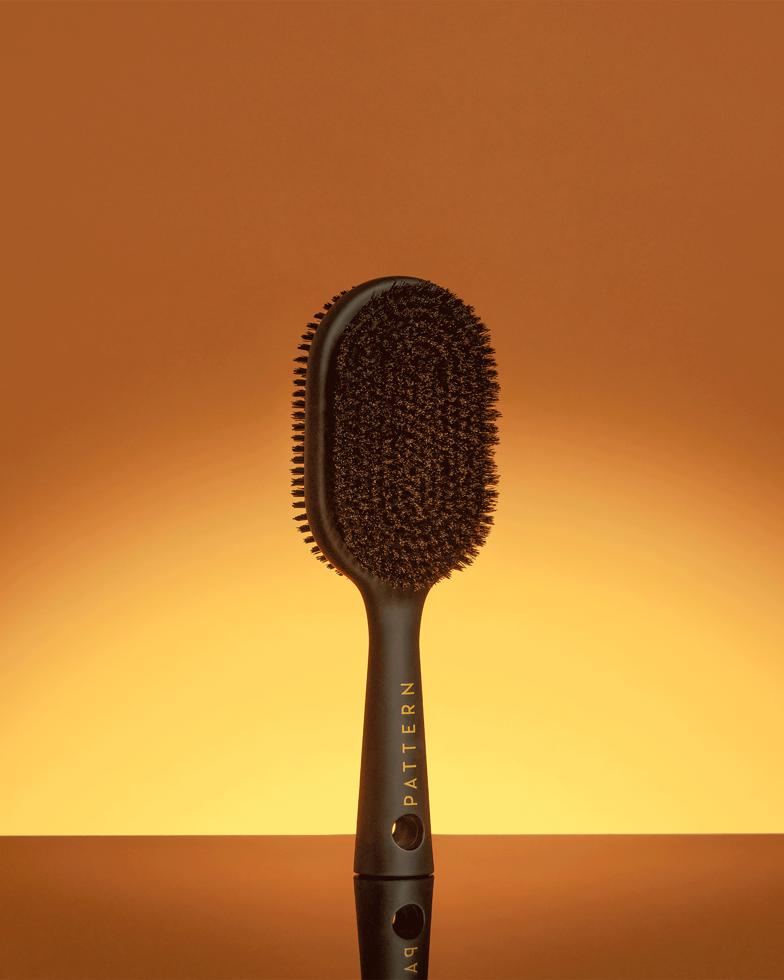 Double-Sided Bristle Brush - PATTERN Beauty by Tracee Ellis Ross double-sided black bristle brush with ergonomic handle against warm gradient background.