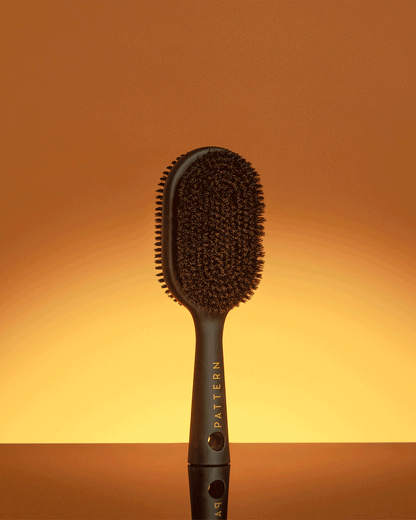 Double-Sided Bristle Brush - PATTERN Beauty by Tracee Ellis Ross double-sided black bristle brush with ergonomic handle against warm gradient background.
