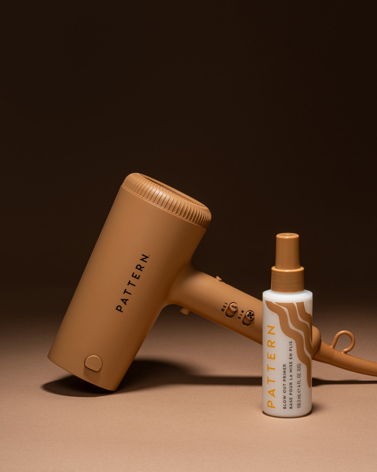 A tan Pattern hair dryer and a matching Pattern hair oil spray bottle, both in a minimalist studio setting with a dark brown background.