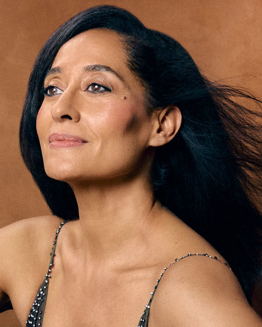 A close-up portrait of a woman with dark hair wearing a beaded black spaghetti-strap top against a warm brown background.