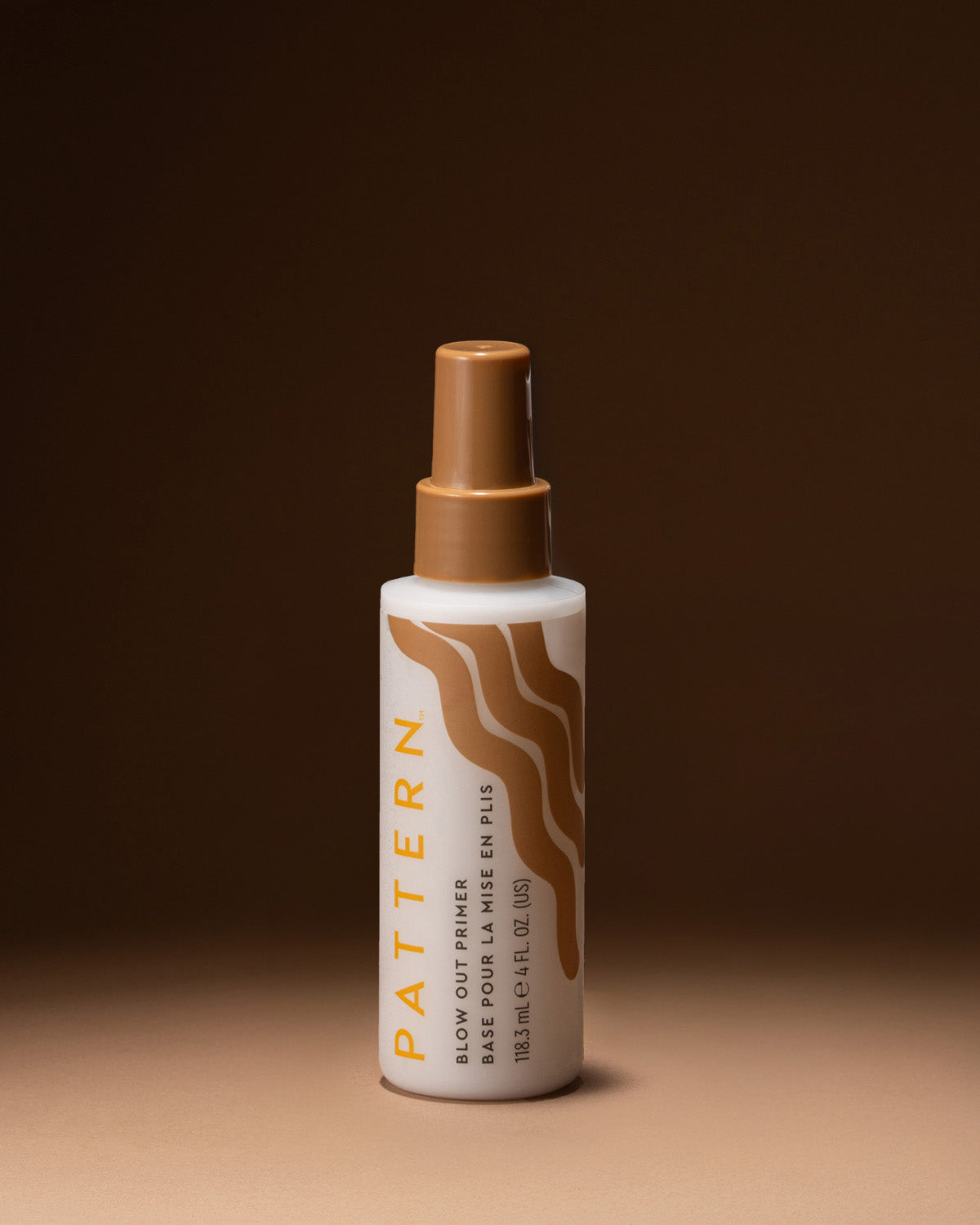 A bottle of Pattern Blow Out Primer & Fin Polish spray, featuring a white bottle with a brown spray nozzle and brown wavy graphic on the label, set against a dark brown background.