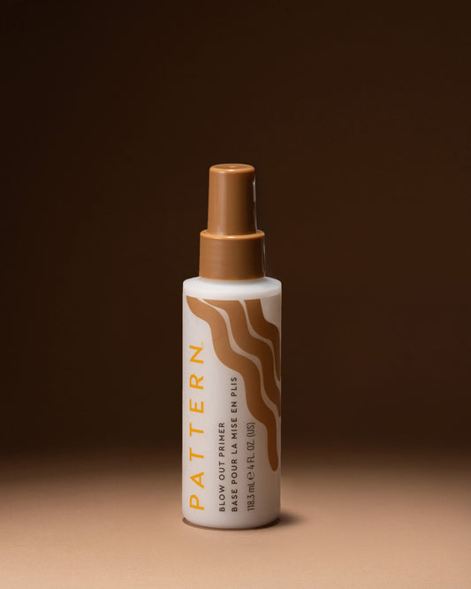 A bottle of Pattern Blow Out Primer & Fin Polish spray, featuring a white bottle with a brown spray nozzle and brown wavy graphic on the label, set against a dark brown background.