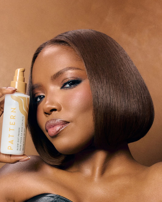 A woman with a sleek, straight brown bob hairstyle holds a bottle of Pattern Hair Oil & Serum against a warm brown studio background.