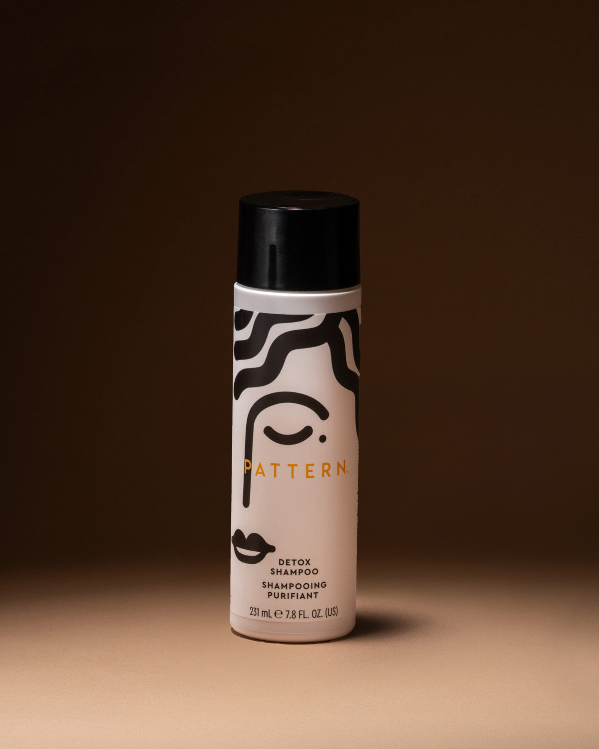 A single can of Pattern Detox Shampoo stands upright on a warm beige surface against a dark brown studio background. The bottle is off-white with a black cap and black graphic line art, with the brand name 'PATTERN' printed in gold/yellow across the front.