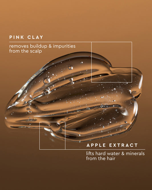 A glossy brown translucent cosmetic smear on a brown gradient background with white text reading 'PINK CLAY' and 'APPLE EXTRACT' plus descriptive claims about scalp and hair buildup.