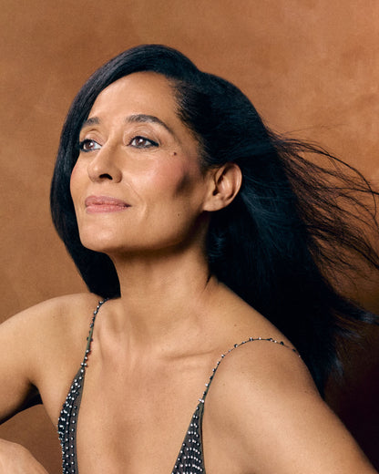 A close-up portrait of a woman with long dark hair wearing a black beaded spaghetti-strap top against a warm brown textured background.