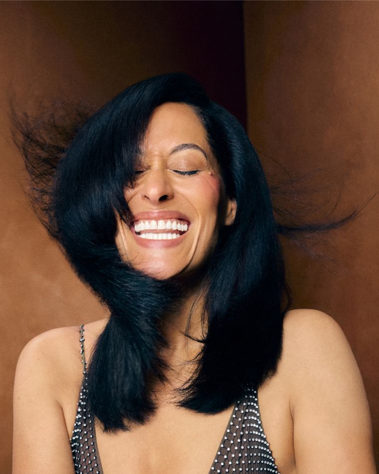 Flexible Hold Spray - Smiling woman with long, glossy black hair wearing a black patterned sleeveless top against a warm brown backdrop.