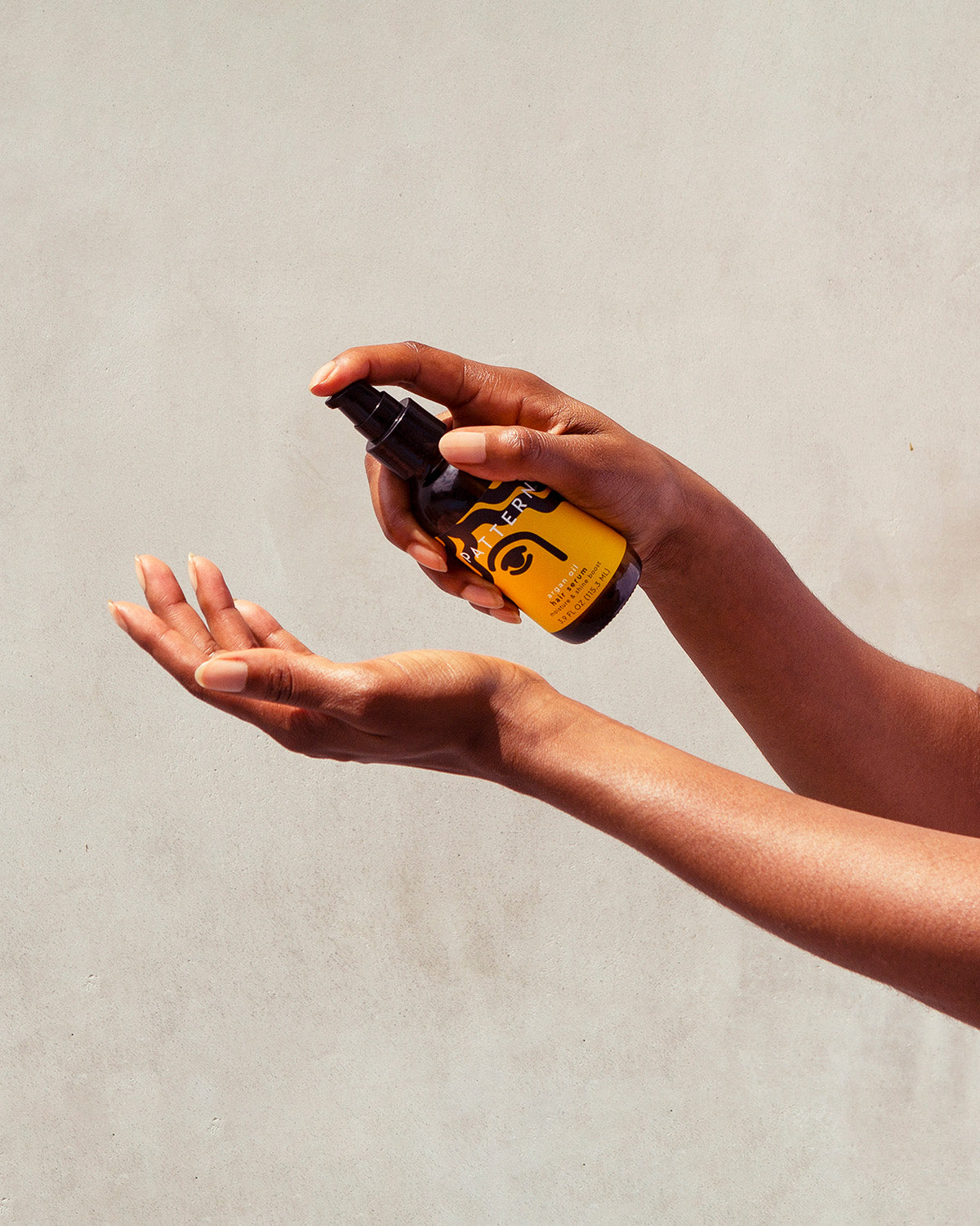 Argan Oil Blend - Amber glass bottle with black pump dispenser labeled PATTERN Beauty Argan Oil Blend held over a pair of open hands against a