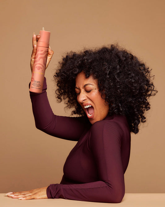 Palo Santo Curl Mousse - PATTERN Beauty Palo Santo Curl Mousse with peach-colored bottle held by a smiling woman with natural curly hair wearing a.