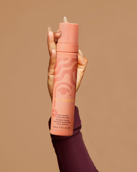 Palo Santo Curl Mousse - Tall cylindrical peach-pink bottle of PATTERN Beauty Palo Santo Curl Mousse with pump dispenser held by hand against beige.