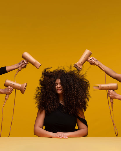 The PATTERN Blow Dryer - Matte peach-colored PATTERN Beauty blow dryers with ergonomic handles around woman with voluminous curly hair on yellow.