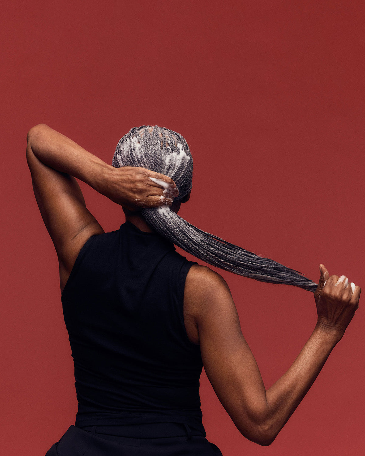 Palo Santo Curl Mousse - PATTERN Beauty Palo Santo Curl Mousse bottle with brown cap held by a woman with long braided hair against a rust-colored.