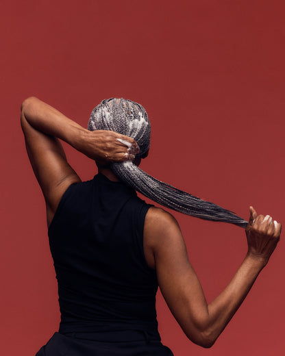 Palo Santo Curl Mousse - PATTERN Beauty Palo Santo Curl Mousse bottle with brown cap held by a woman with long braided hair against a rust-colored.