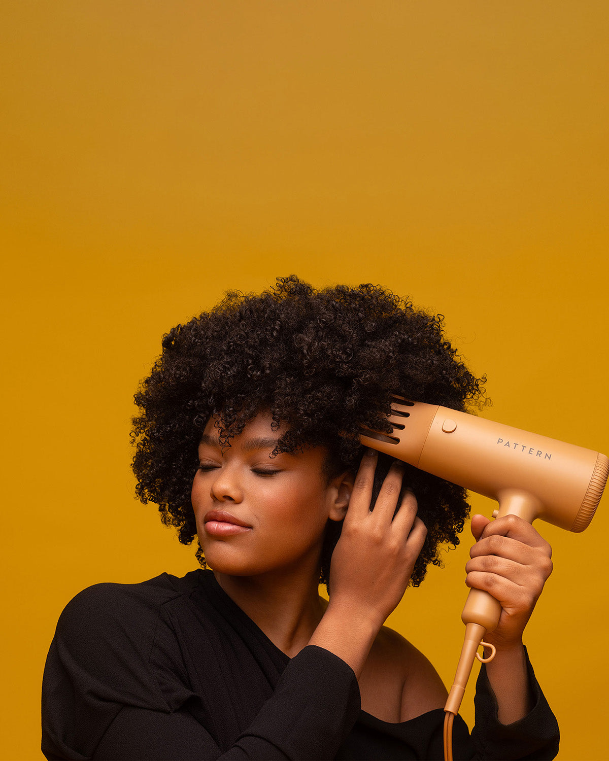The PATTERN Blow Dryer - Matte peach-colored PATTERN Beauty blow dryer with rounded edges held near curly hair against a mustard backdrop.