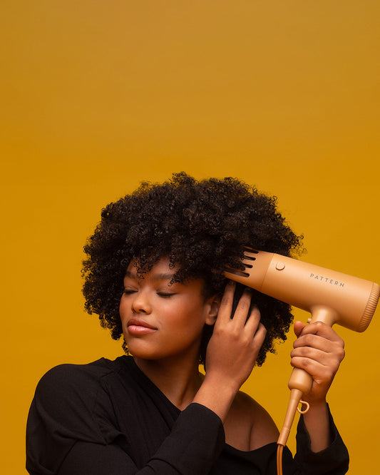The PATTERN Blow Dryer - Matte peach-colored PATTERN Beauty blow dryer with rounded edges held near curly hair against a mustard backdrop.