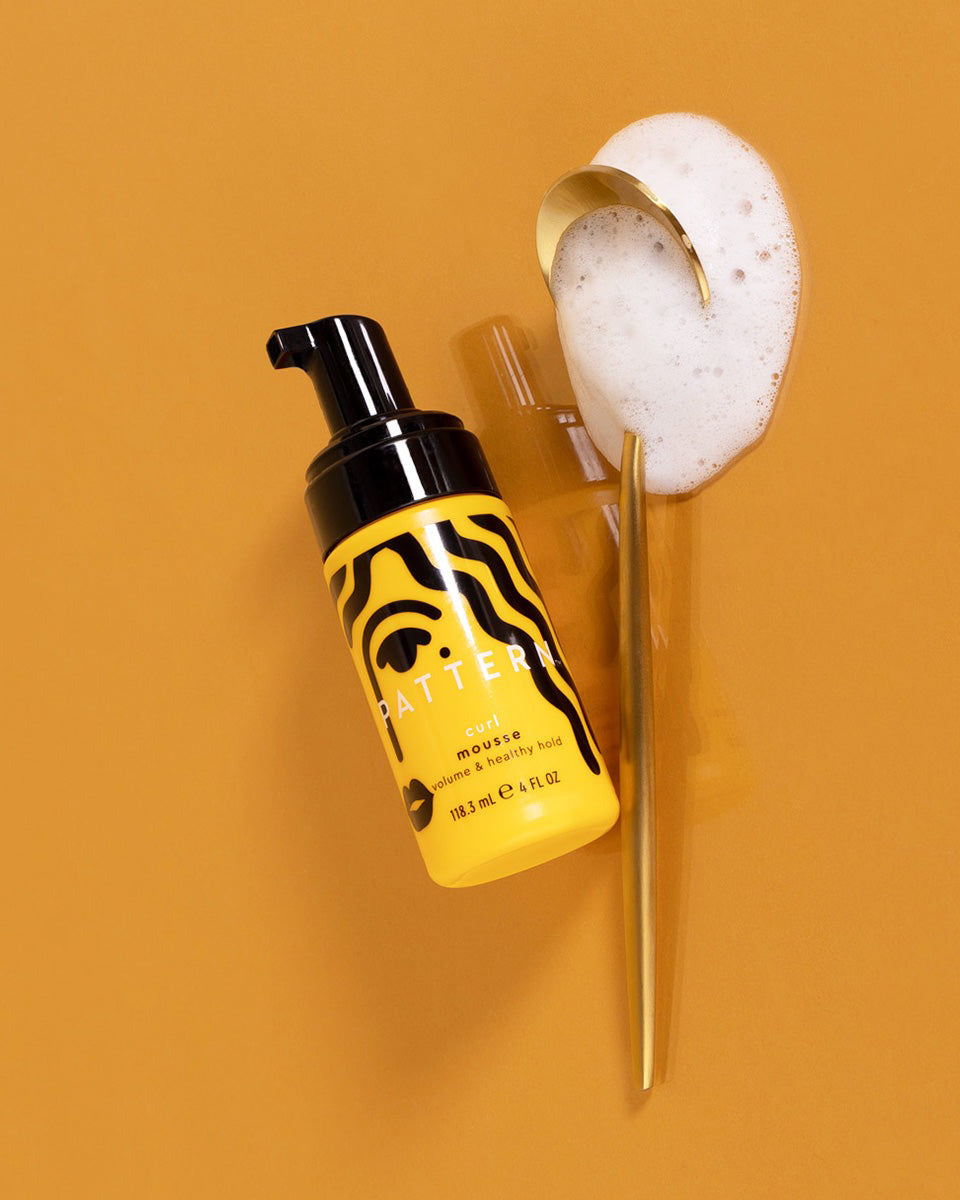 Curl Mousse - Yellow bottle of PATTERN Beauty Curl Mousse with black tiger stripes and foam on transparent applicator wand against mustard