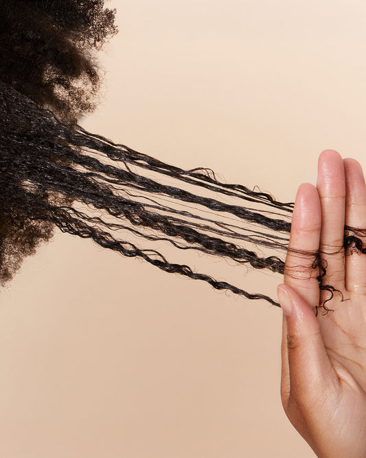 Breakage Barrier Hair Mask - Four strands of shiny, black, tightly coiled hair held between fingers against a neutral background.