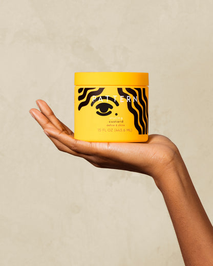 Styling Custard - Hand holding yellow jar of PATTERN Beauty by Tracee Ellis Ross Styling Custard with black abstract hair design.